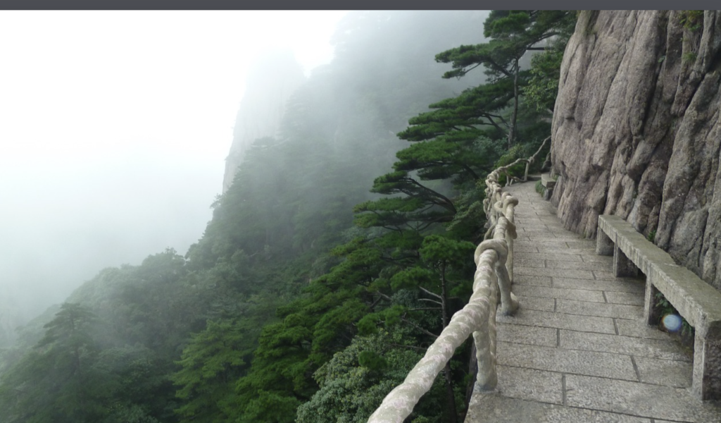 The image depicts a tranquil, misty mountain landscape, with a winding stone path carved into the cliffside. Tall, ancient pine trees line the path, creating a sense of timeless serenity. The atmosphere is shrouded in fog, adding an air of mystery and introspection to the scene. This visually striking setting could serve as a powerful metaphor for the journey of overcoming career setbacks, as described in the accompanying text. The image evokes a sense of challenge and determination, with the path leading the viewer onward, despite the obscuring conditions. This visual representation aligns well with the themes of resilience, growth, and self-discovery discussed in the document.